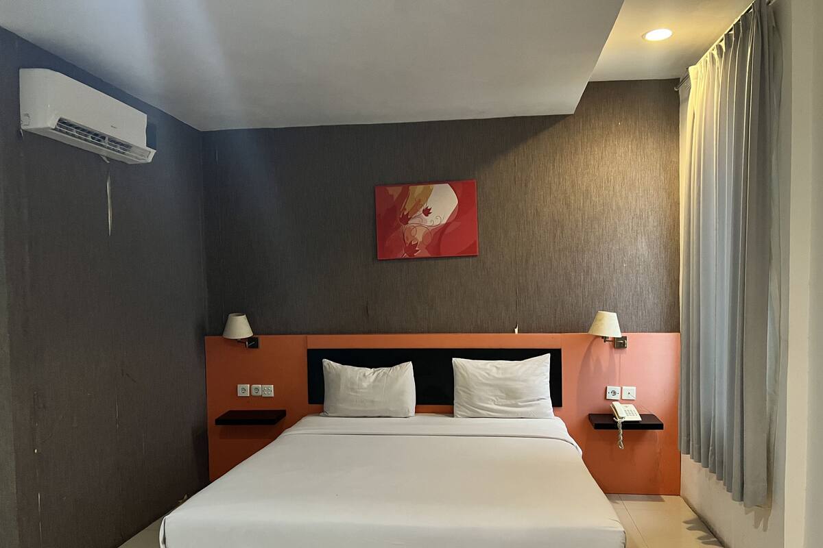 A hotel room with a large bed, two bedside tables with lamps, an air conditioning unit, a painting on the wall, and a curtain on the window.