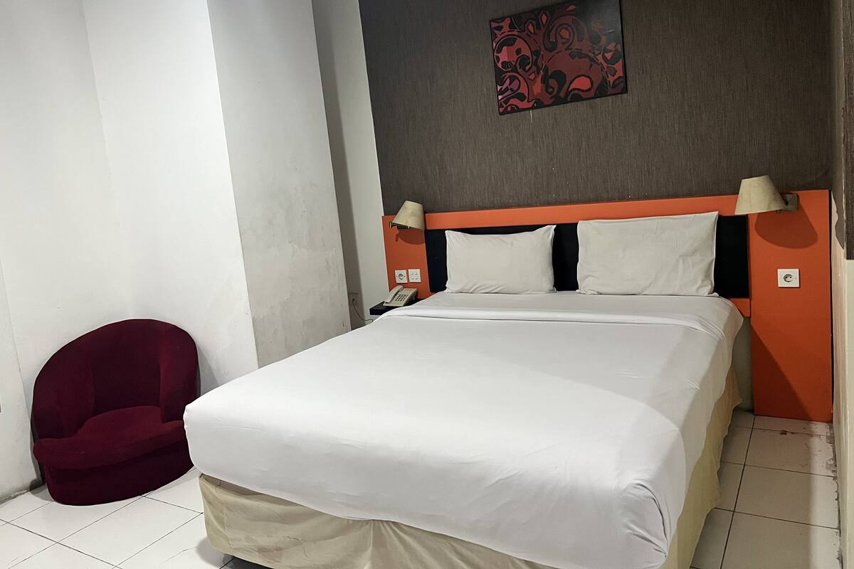 A hotel room with a bed, a red chair, a telephone, and a wall-mounted artwork.