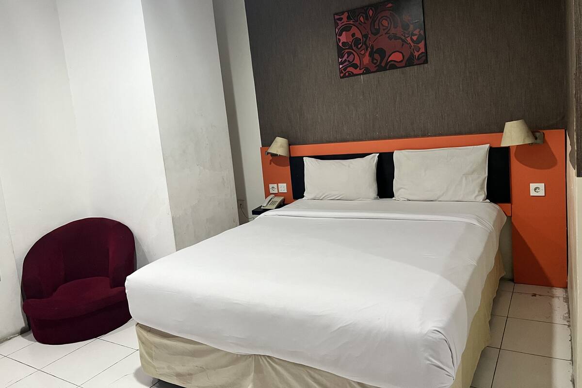 A hotel room with a bed, a red chair, a telephone, and a wall-mounted lamp.