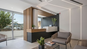 A modern reception area with a wooden reception desk, a seating area with a sofa and armchairs, a coffee table with a plant and books, and a large window offering a view of the city. - Ostrakon Suites (Agia Marina)