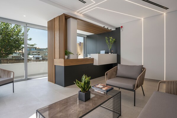 A modern reception area with a wooden reception desk, a seating area with a sofa and armchairs, a coffee table with a plant and books, and a large window offering a view of the city. - Ostrakon Suites (Agia Marina)
