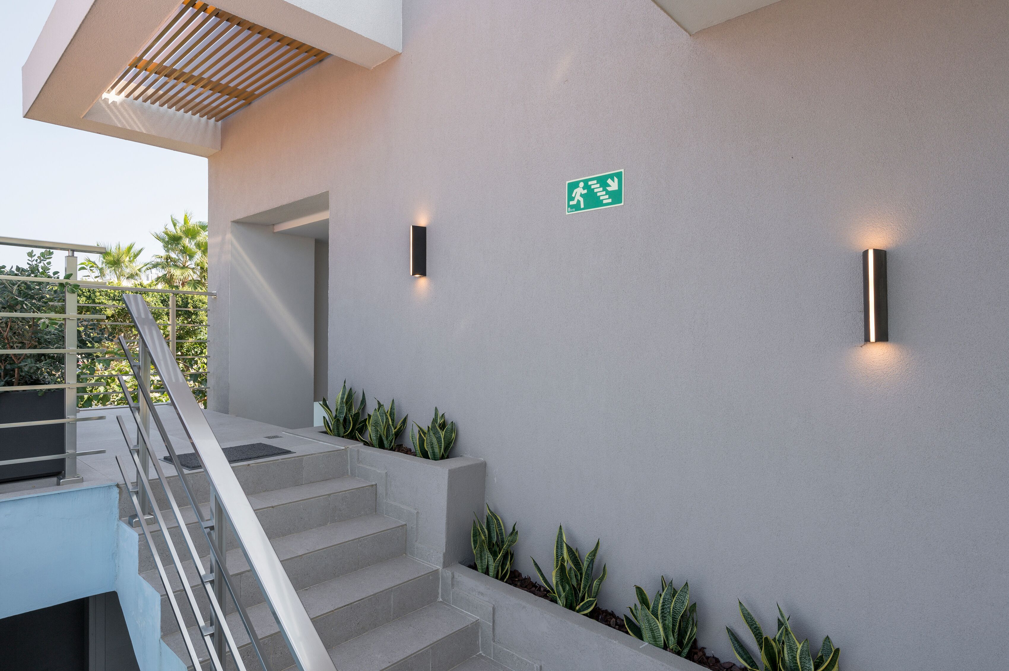 Una escalera moderna con barandilla de metal, un letrero de salida de emergencia verde y luces fijadas a la pared.