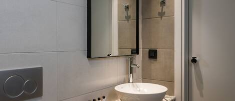 A modern bathroom with a white sink, a mirror, and a wall-mounted faucet. There are three bottles of toiletries on a wooden shelf.