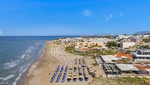 A coastal beach area with rows of umbrellas and beach chairs, a promenade, and buildings with balconies overlooking the beach. - Ostrakon Suites (Agia Marina)