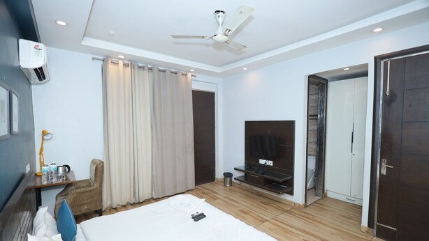 A hotel room with a bed, a TV, a ceiling fan, an air conditioning unit, a desk with a lamp, and a door leading to another room. - Wedlock Hotels (Gurugram)