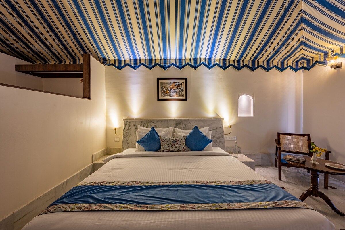 A hotel room with a striped ceiling, a bed with blue pillows, a framed picture on the wall, and a small table with a chair.