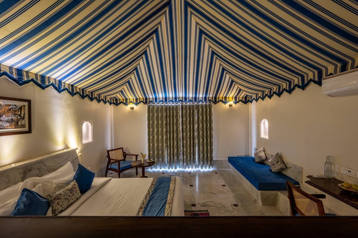 Ein Hotelzimmer mit gestreifter Decke, einem Bett mit blauer Bettwäsche, einer Couch, einem Sessel und einem Vorhang mit vertikalen Streifen.