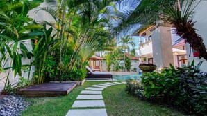 A lush garden with a pool, stone pathway, and a wooden deck with a lounge chair. - Cempaka Villa by Premier Hospitality (Nusa Dua)