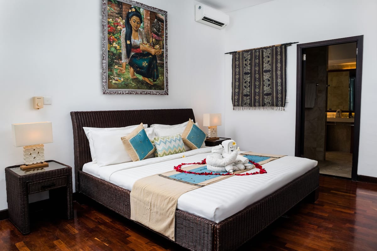 A spacious bedroom with a large bed, two bedside tables, a wall-mounted air conditioner, a decorative painting, and a patterned curtain.