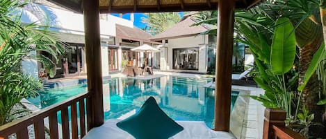 Vista de la piscina con una cama, sillas de descanso y plantas tropicales.