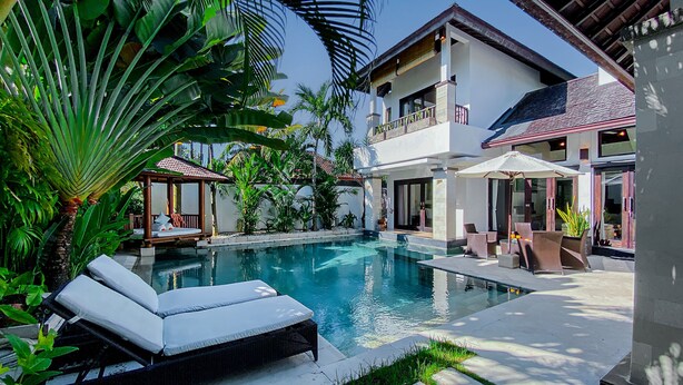 A pool area with lounge chairs, a covered seating area, and a two-story building with balconies. - Cempaka Villa by Premier Hospitality (Nusa Dua)