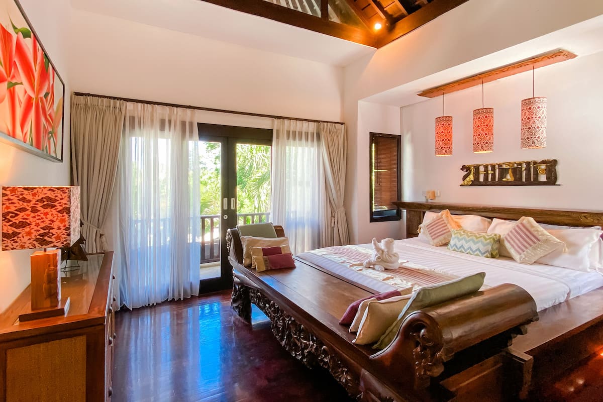 A spacious bedroom with a large bed, a wooden nightstand, a desk with a lamp, and a view of greenery through the window.