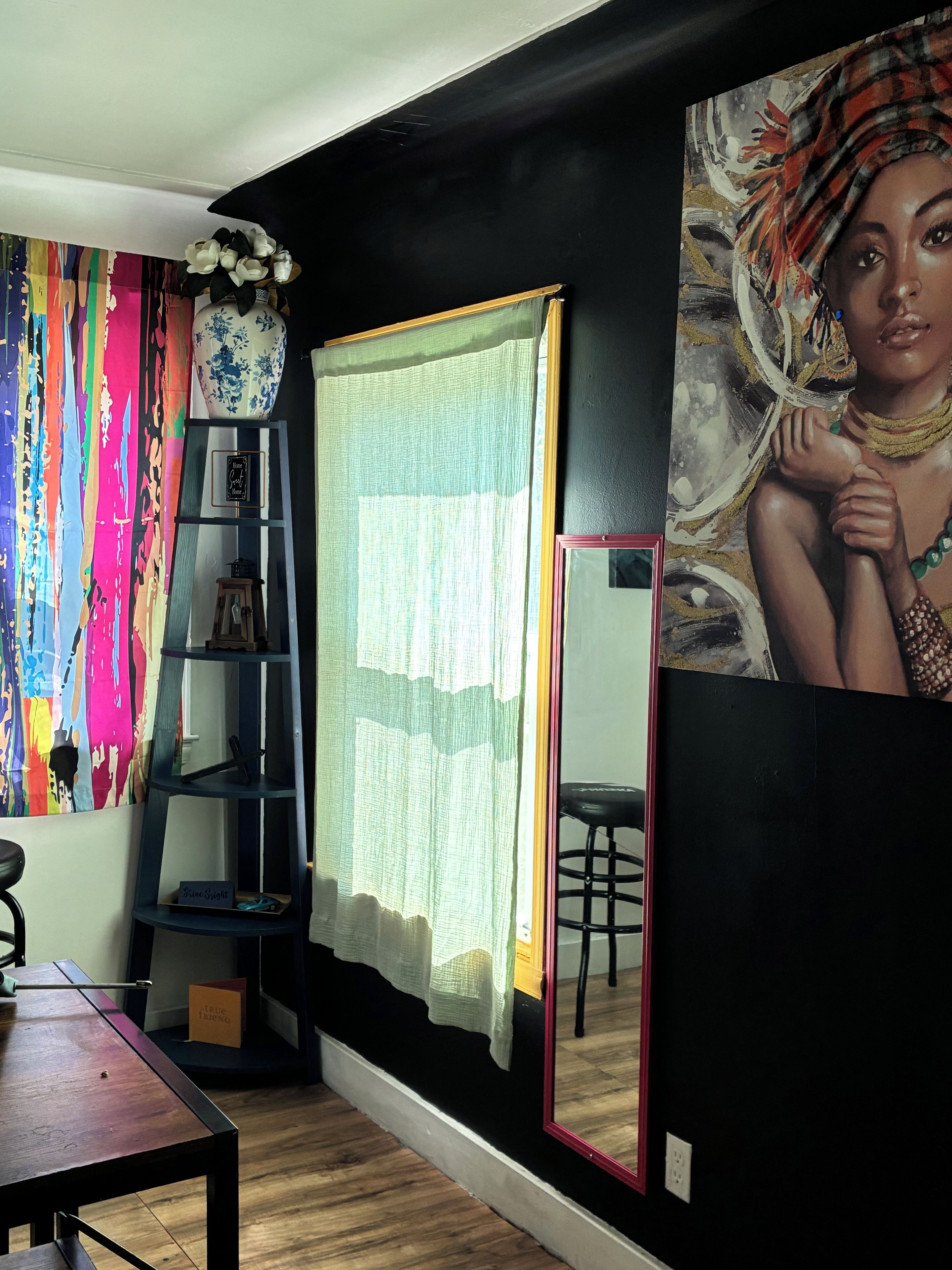 A room with a wooden floor, a black wall, a colorful curtain, a white vase with flowers, a blue shelf with items, and a large painting.