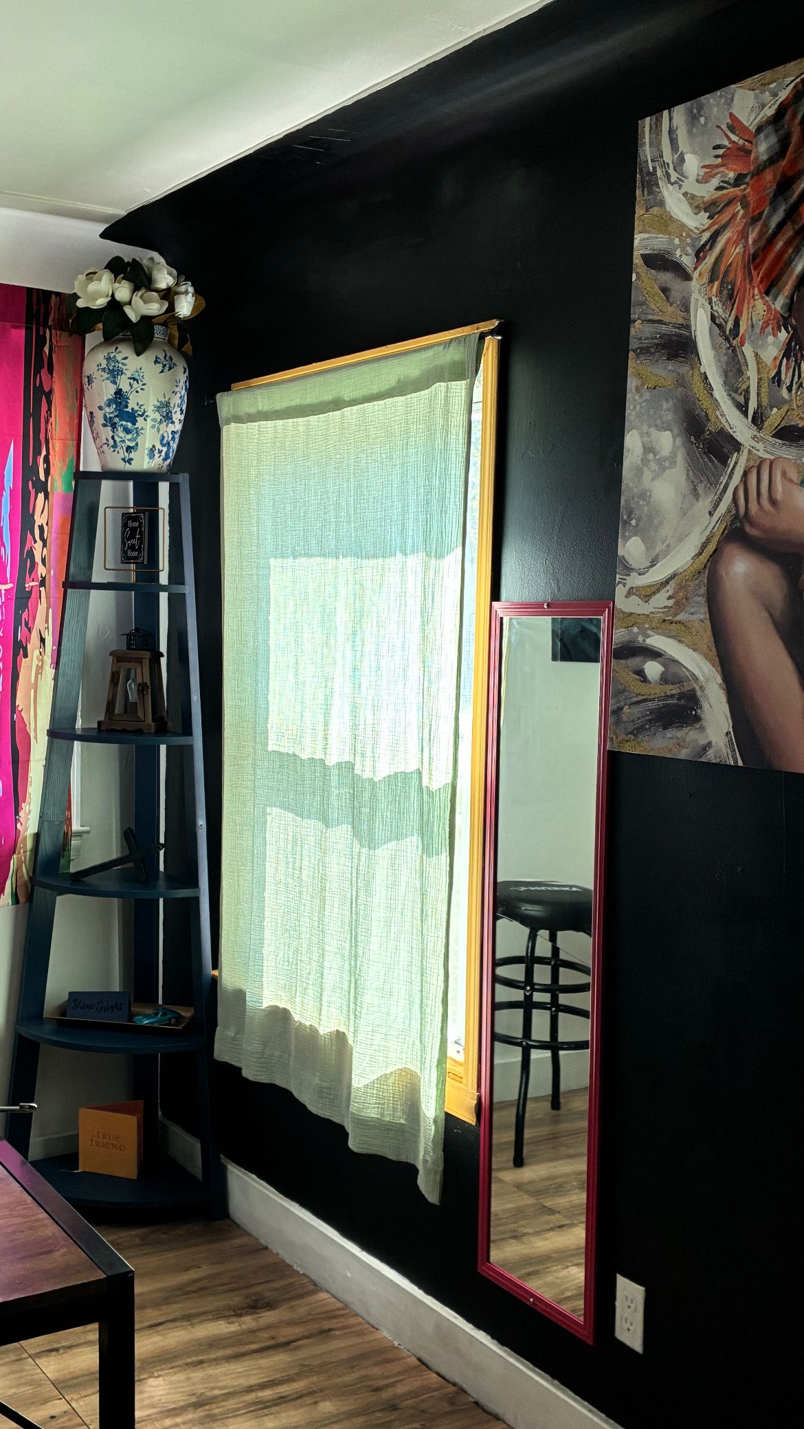 Une pièce avec un mur noir, une fenêtre ornée d’un rideau léger, un miroir, une échelle en bois et une étagère sur laquelle sont disposés des objets décoratifs.