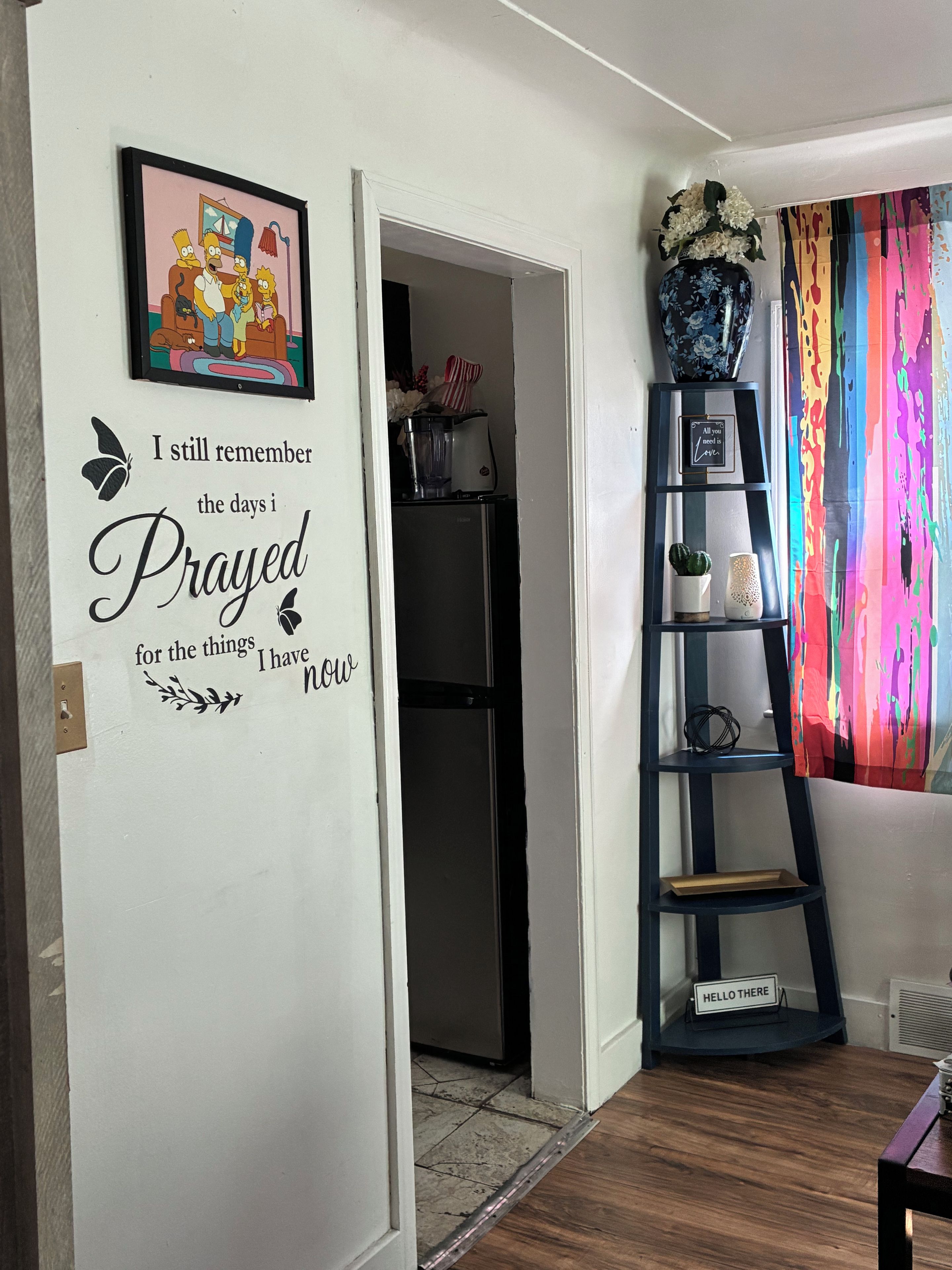 A room with a wall decal featuring a quote, a framed picture, a shelf with decorative items, and a colorful curtain.
