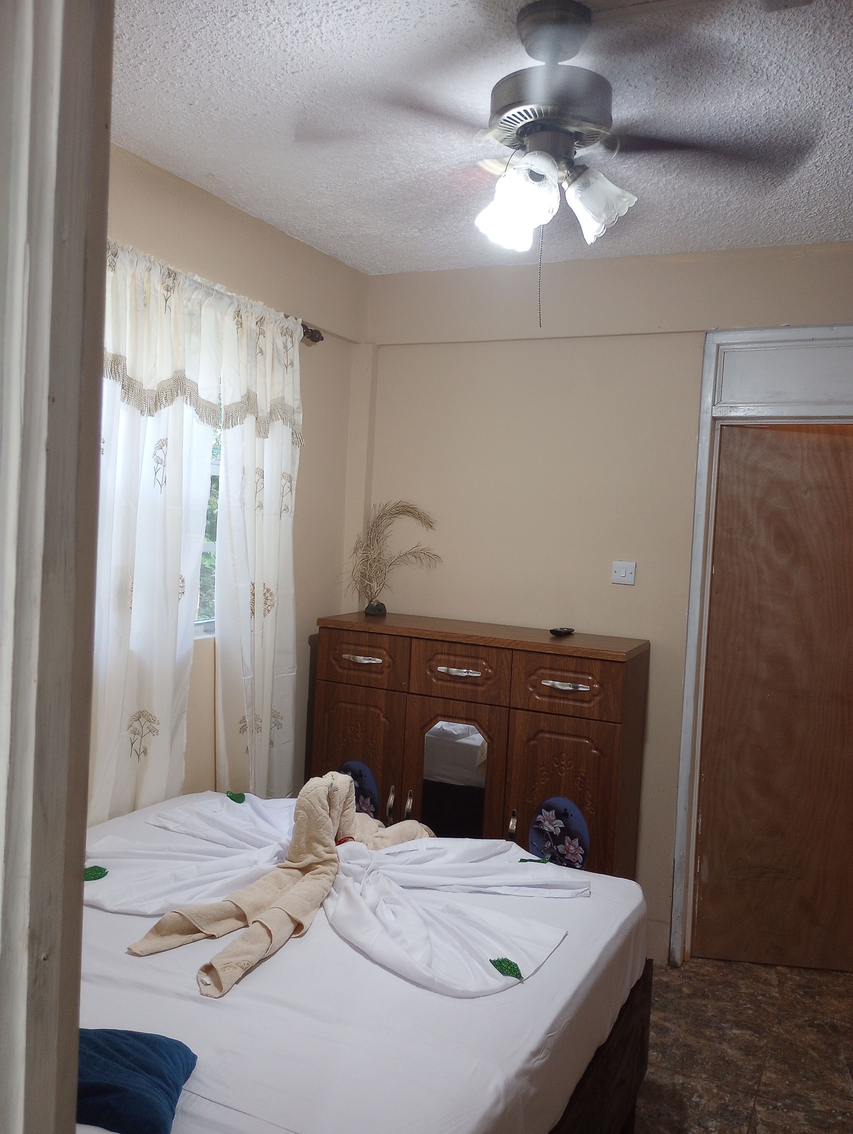 Une chambre à coucher avec un lit, une commode en bois, un ventilateur de plafond et une fenêtre avec des rideaux.
