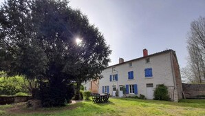 Ein zweigeschossiges Haus mit weißer Fassade und blauen Fensterläden, ein großer Baum im Vorgarten und ein Tisch mit Stühlen draußen.