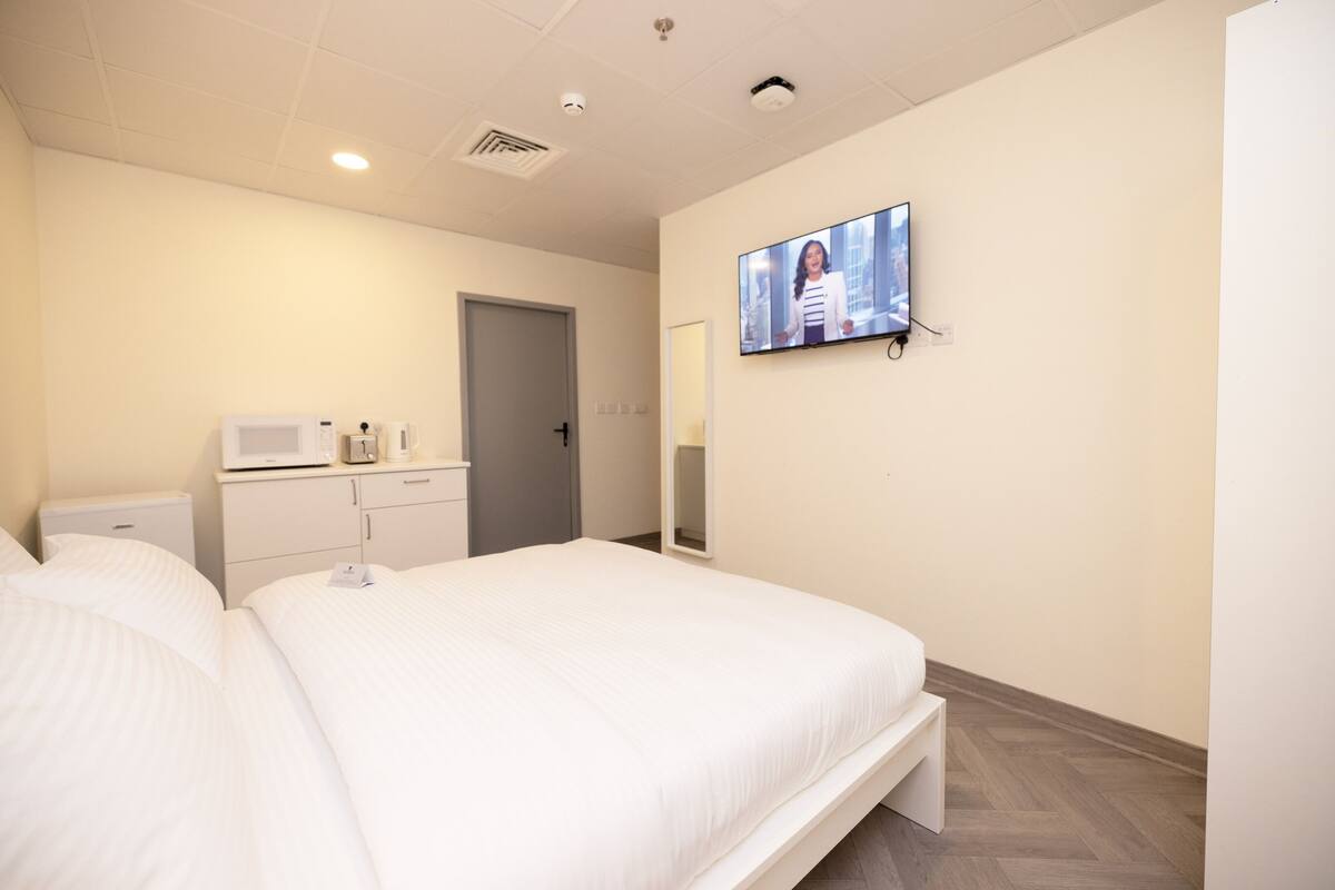 Habitación de hotel con una cama grande, un televisor montado en la pared, un microondas, un mueble pequeño y una puerta.