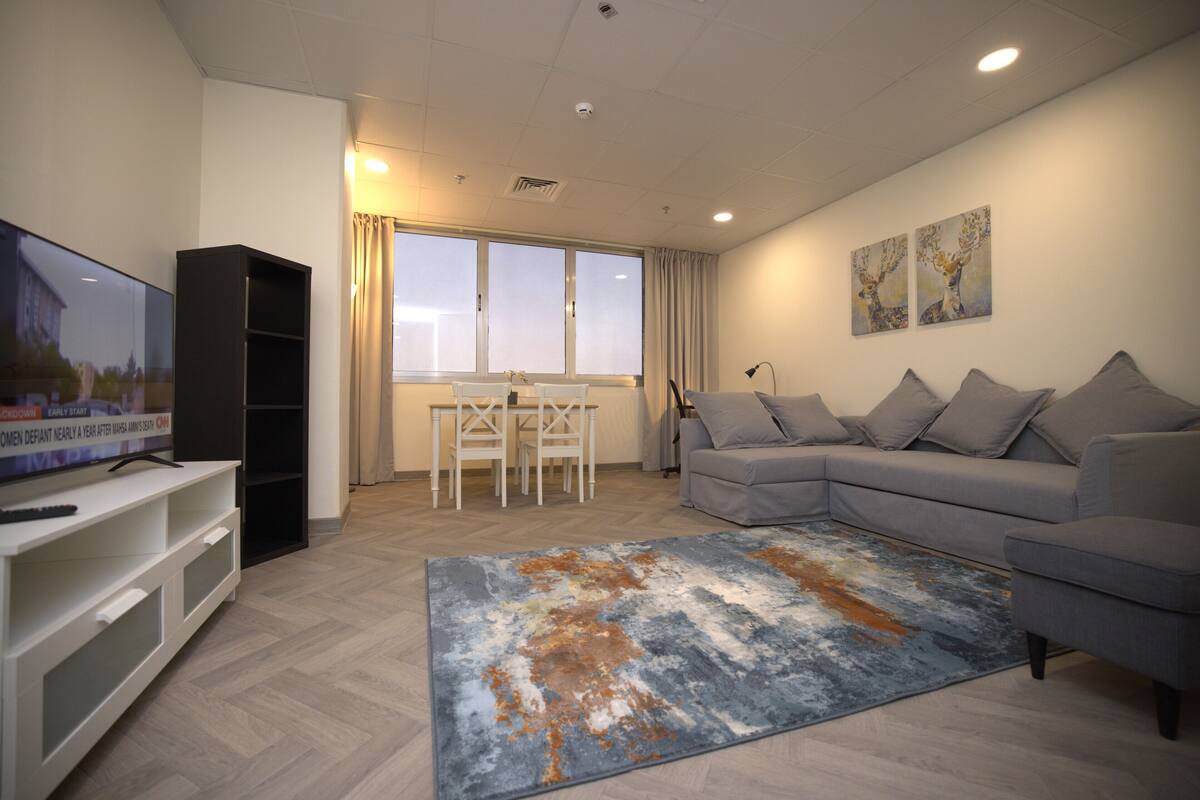 Una sala de estar moderna con un sofá gris, un televisor de pantalla plana, un mueble blanco para el equipo multimedia, una mesa de comedor con sillas y una alfombra grande.