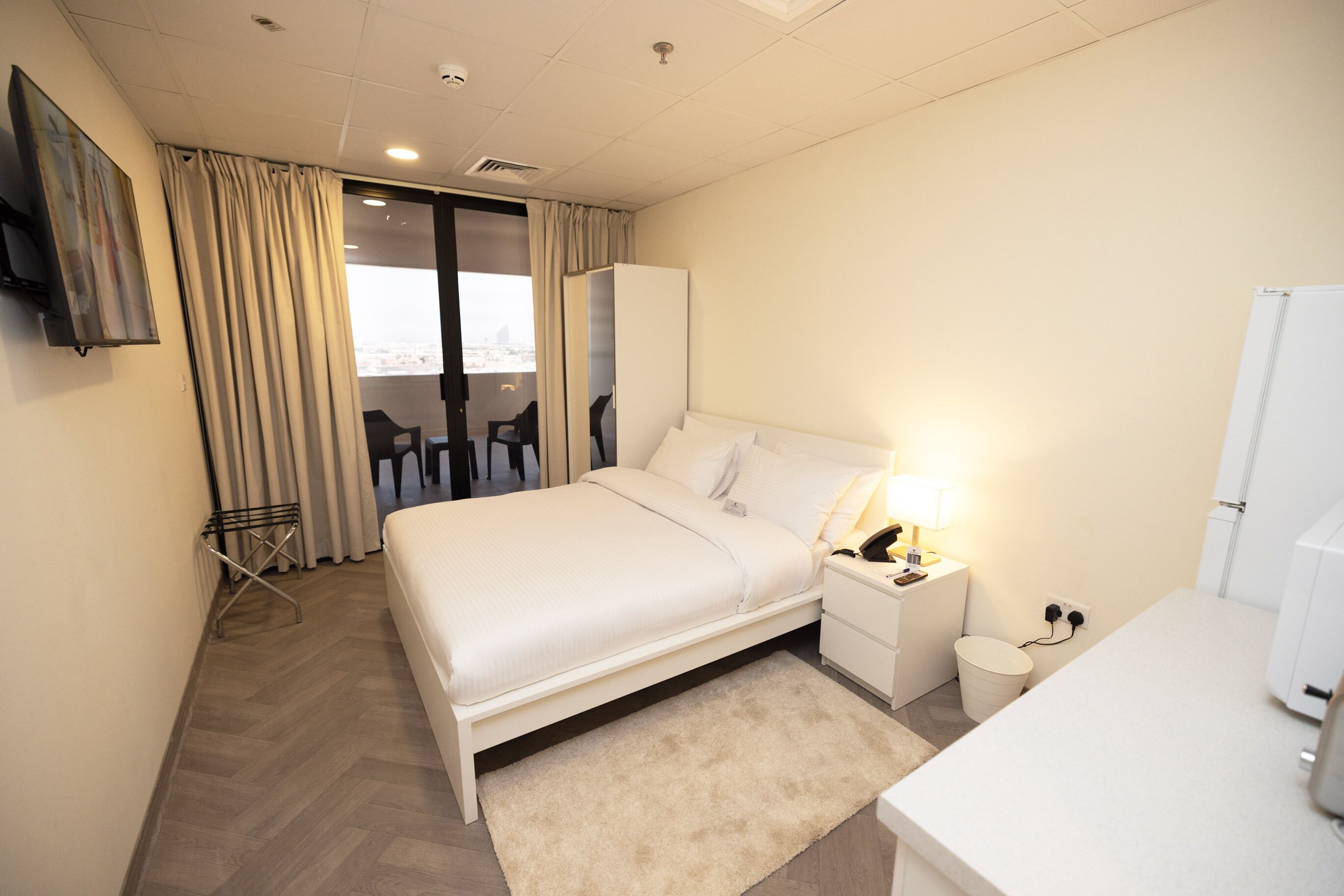 A hotel room with a bed, bedside table, lamp, and a view of the city through the window.