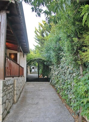 Un sendero estrecho que lleva a una entrada con portón, con un muro de piedra a un lado y abundante vegetación al otro.