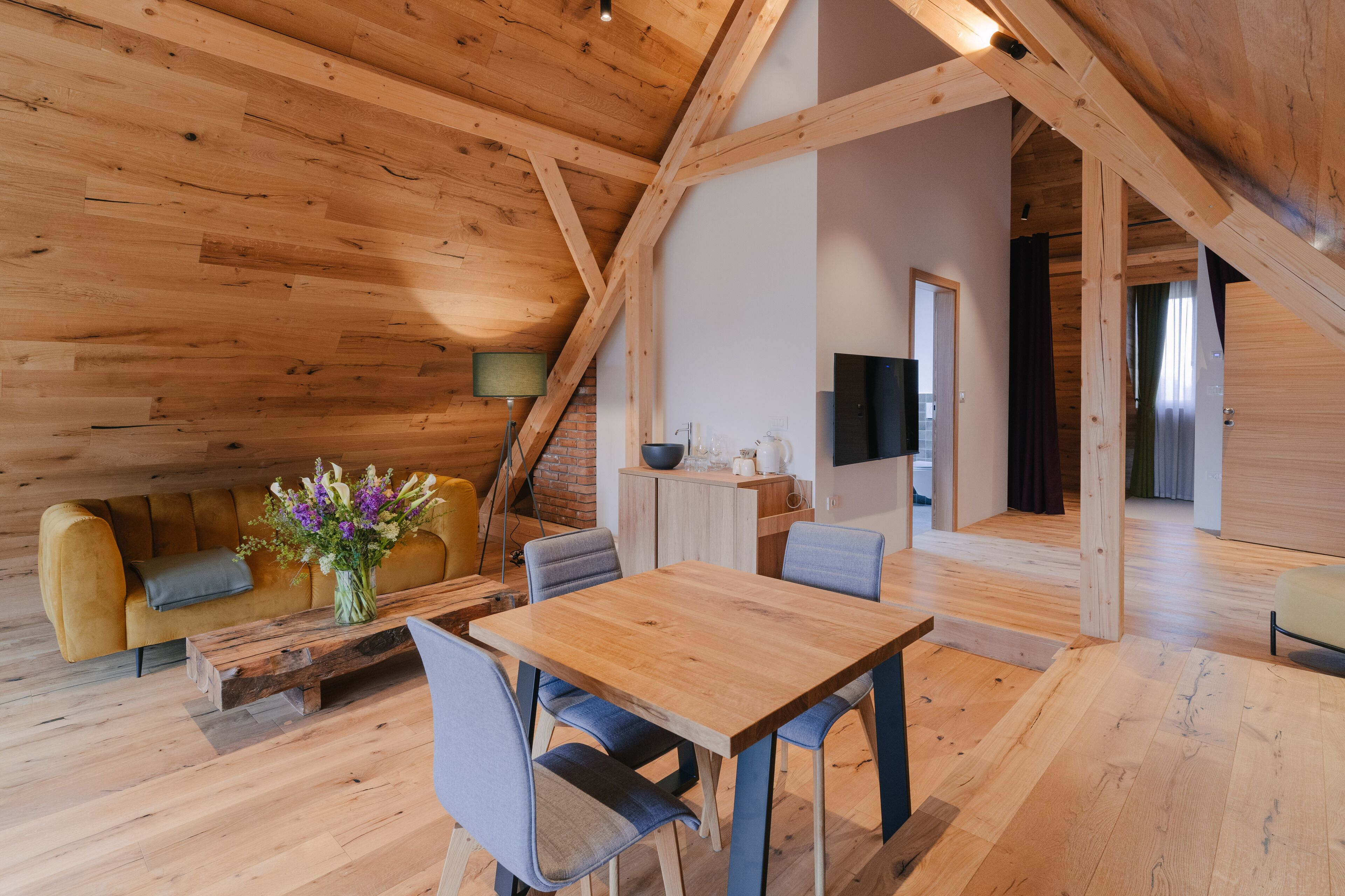 A spacious loft with a high ceiling, wooden beams, a dining area with a wooden table and chairs, a sofa, and a television in the background.