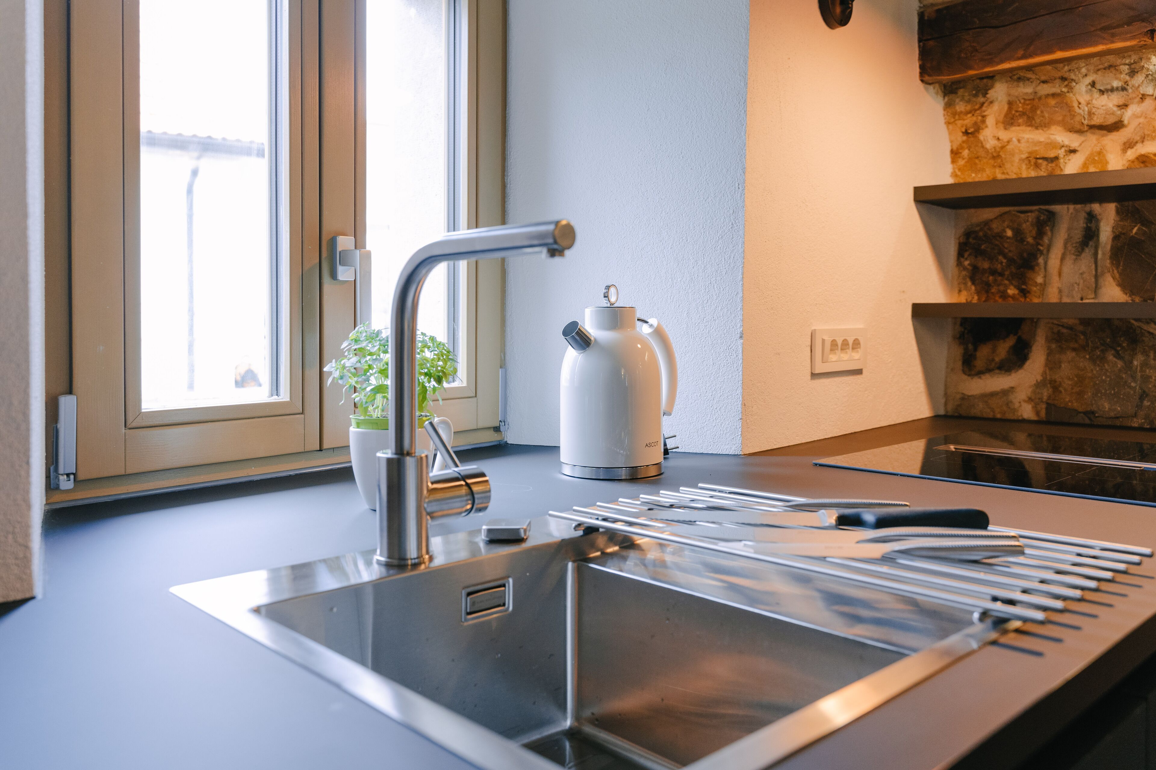 A modern kitchen with a stainless steel sink, a kettle, and a window with a view of the outside.
