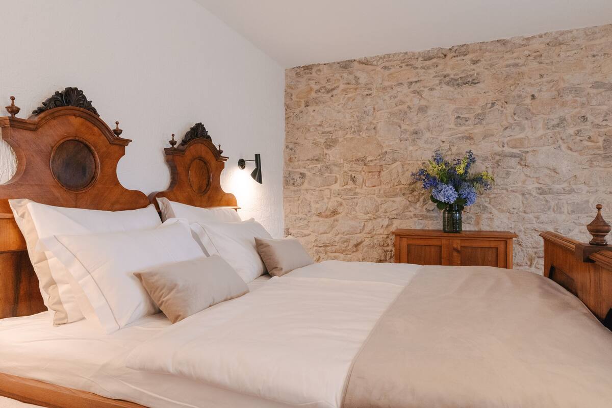 A bed with a wooden headboard, a stone wall, and a nightstand with a vase of flowers.