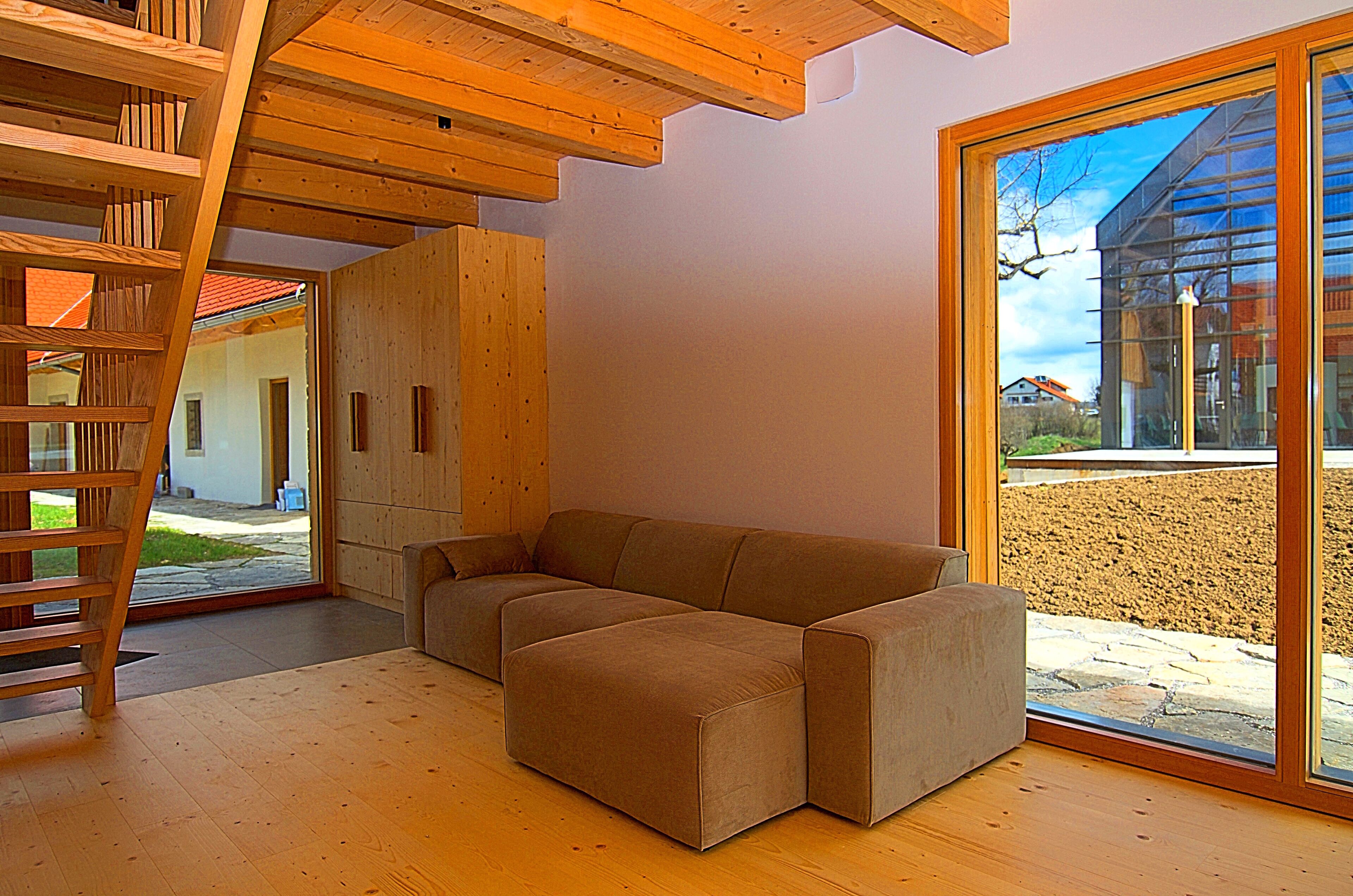 A modern interior with a wooden staircase, a sectional sofa, and a large window offering a view of a building and a gravel path.