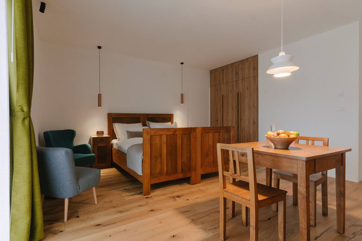 A bedroom with a wooden bed, a green armchair, a wooden dining table with chairs, and pendant lights.