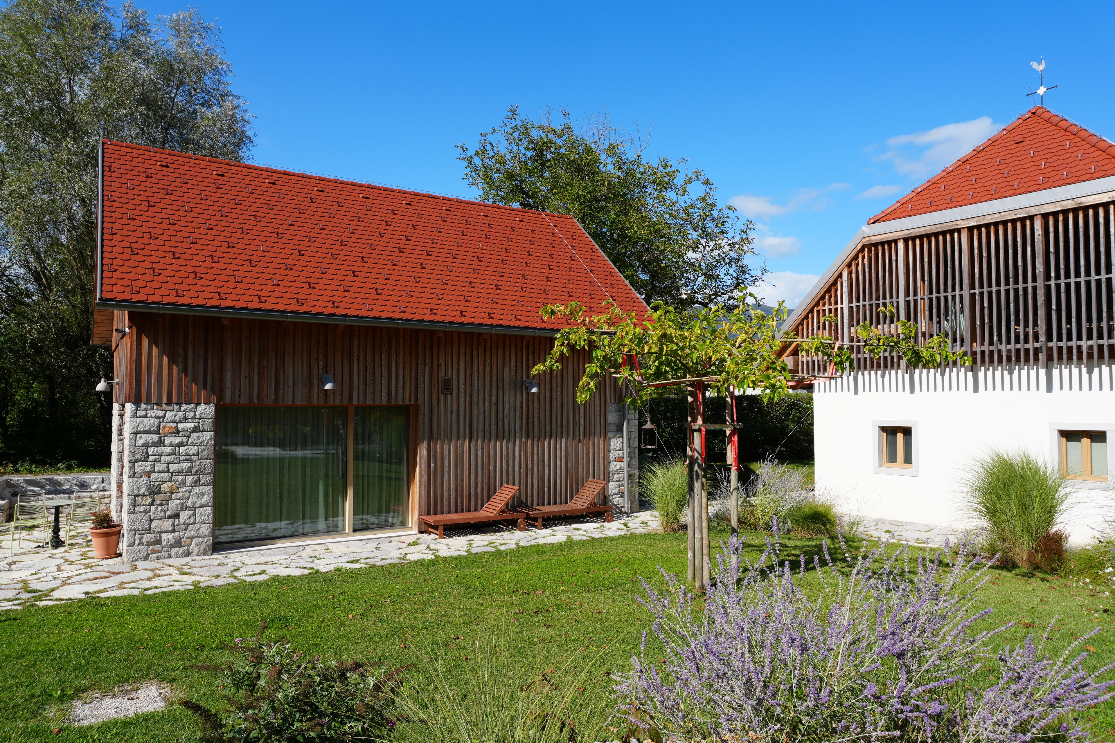 Ett modernt hus med ett rött tegeltak, en stenmur och en träveranda med två solstolar.