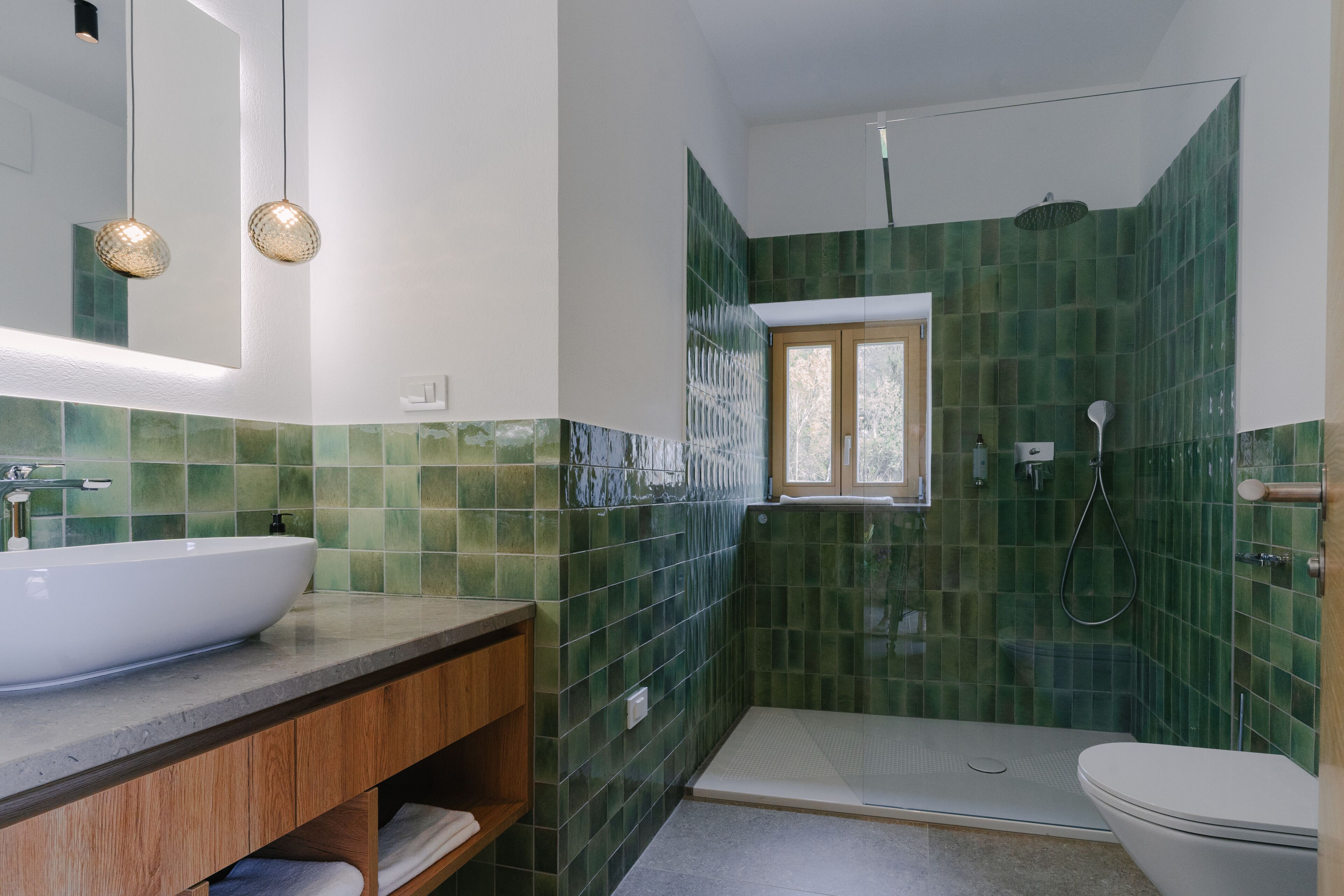 A bathroom with a white sink, wooden vanity, green tiled walls, a window, and a walk-in shower.