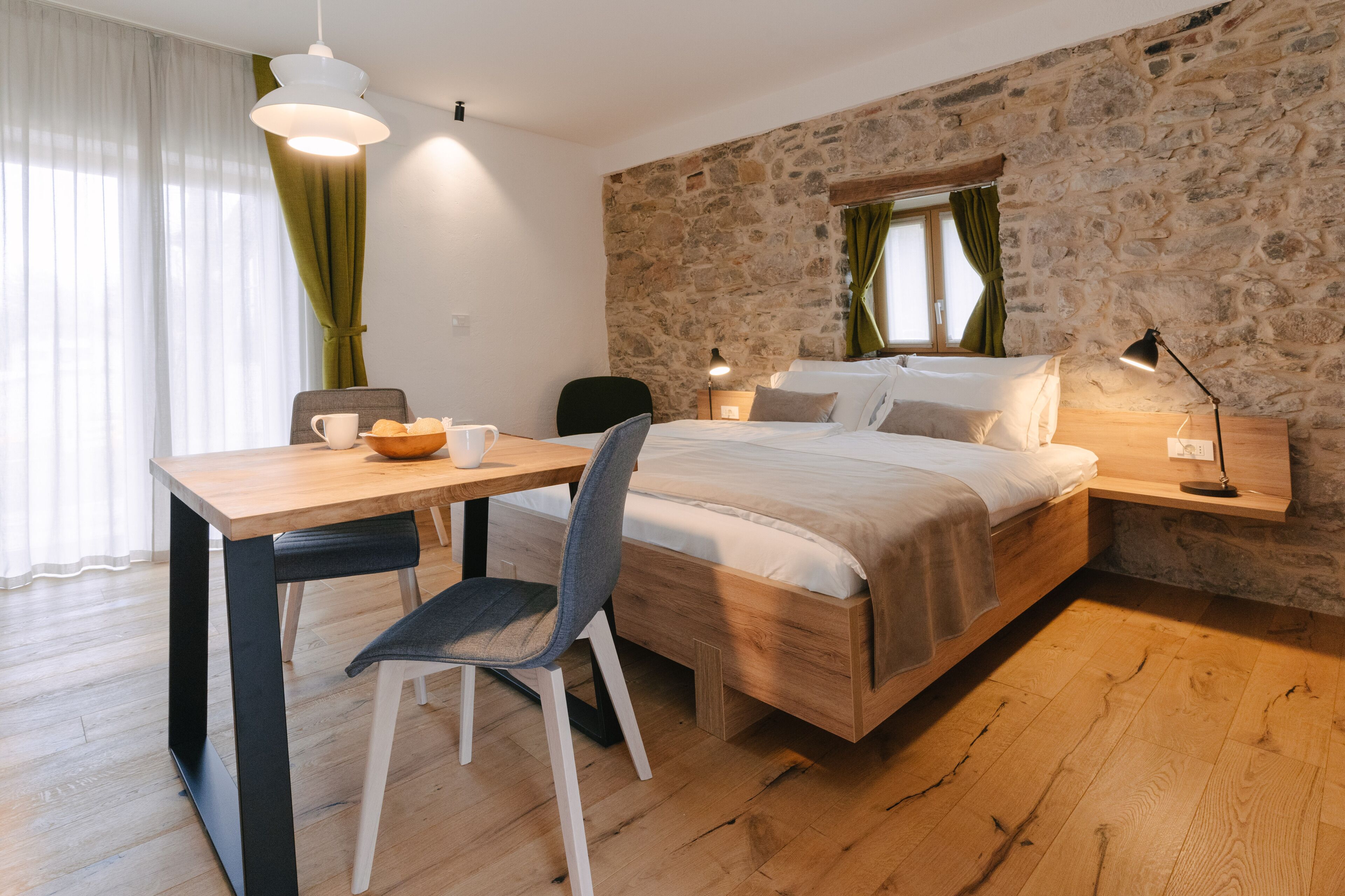 A modern bedroom with a stone wall, a wooden bed, a desk with a chair, and a window with curtains.