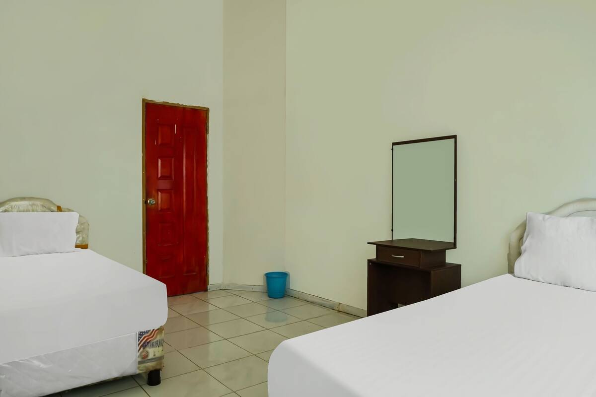 A hotel room with two beds, a red door, a small blue trash bin, and a mirror on a dresser.