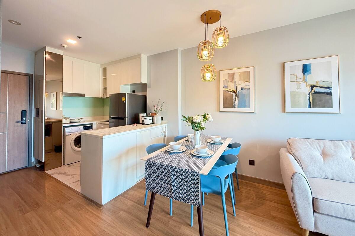 A modern kitchen with a central island, a dining area with blue chairs, and a living space with a sofa.