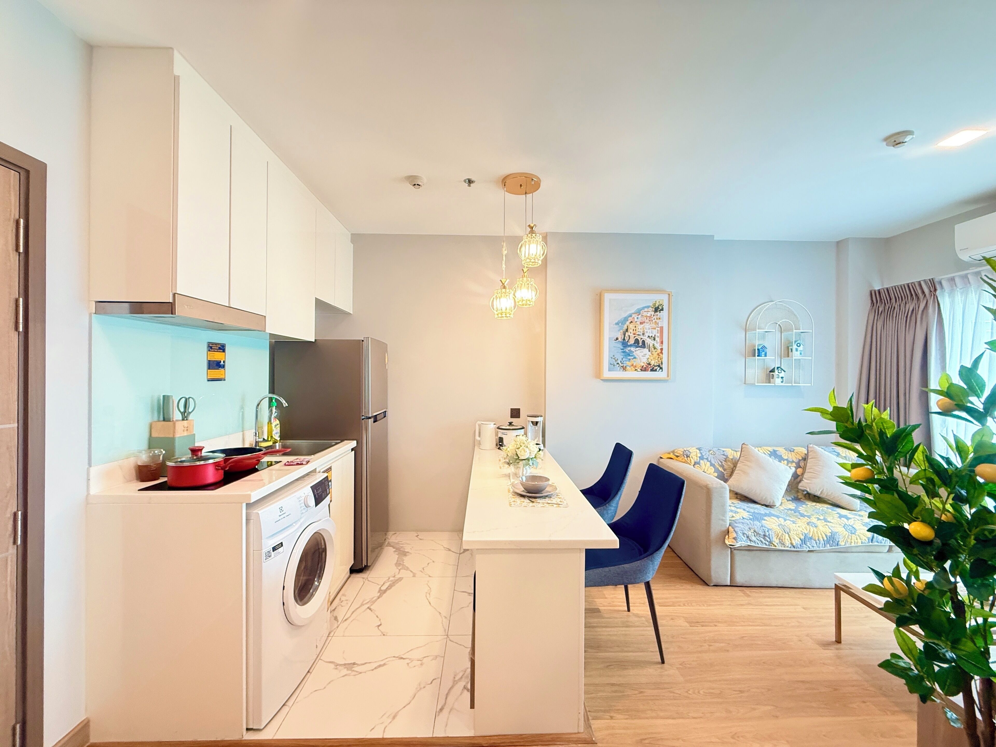 Een moderne keuken met een ingebouwde wasmachine, een eethoek met een centraal eiland, en een woonkamer met een bank en een plant.