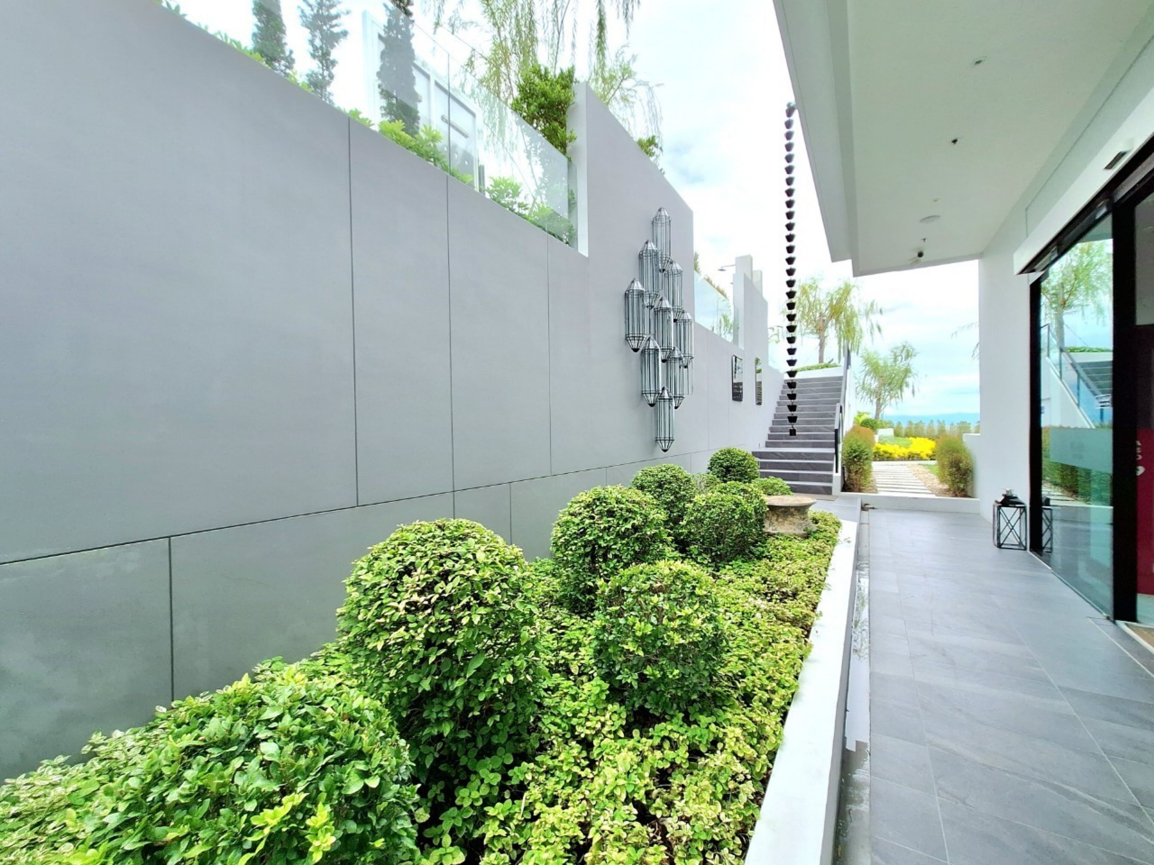 A modern outdoor area with a grey wall, green bushes, and a staircase leading to a higher level.