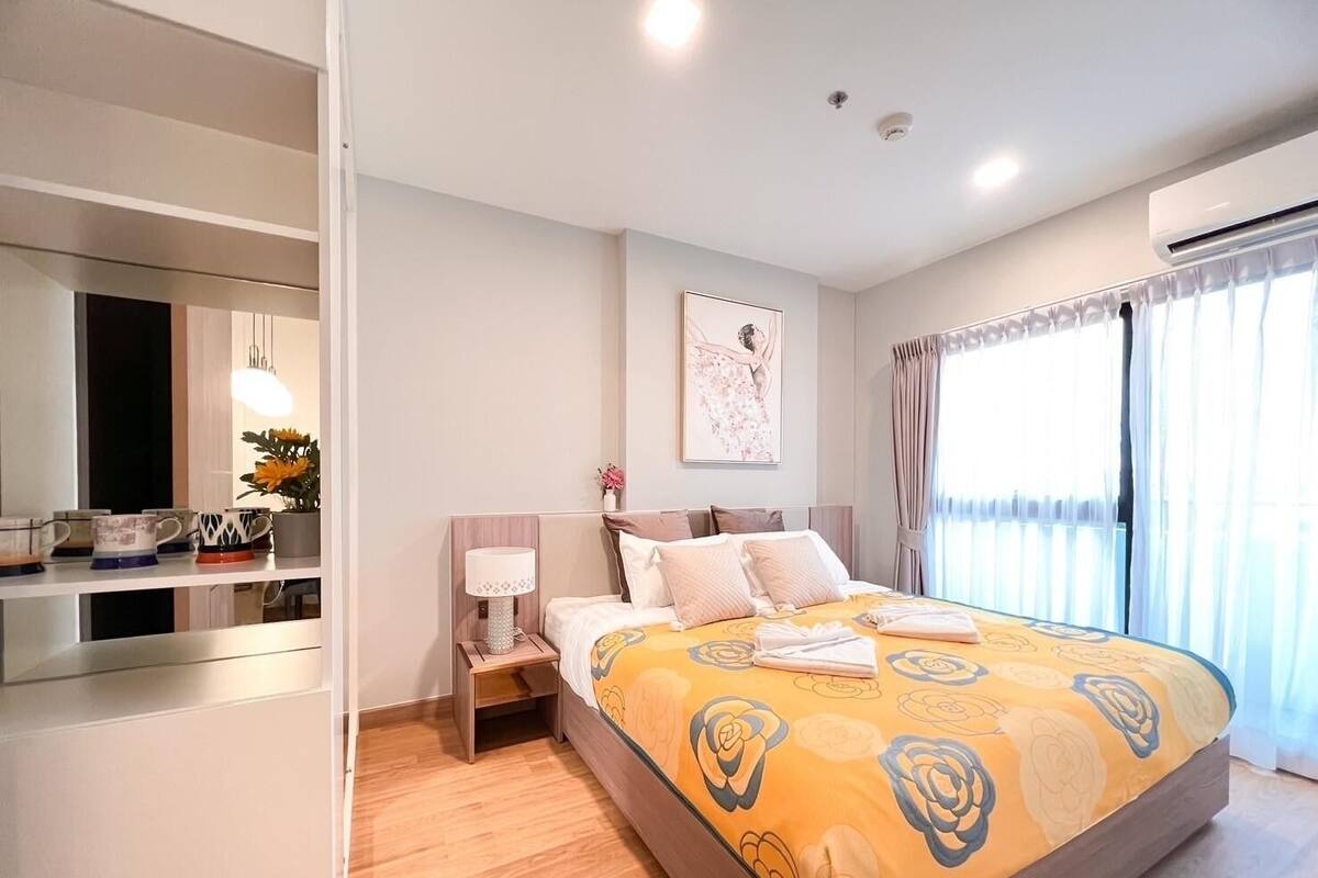 A modern hotel room with a large bed, a bedside table with a lamp, a mirror, and a window with curtains.