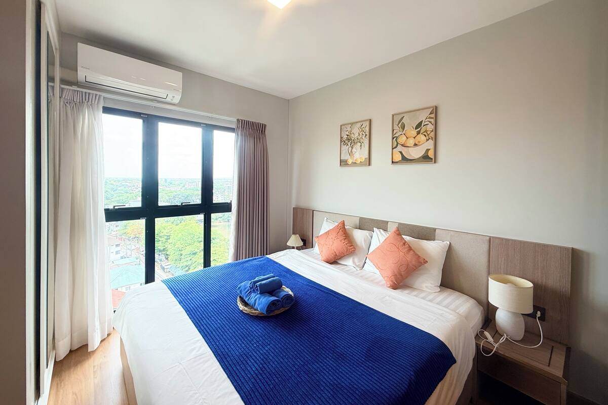 A hotel room with a large bed, a window with curtains, an air conditioning unit, a bedside table with a lamp, and two wall-mounted paintings.