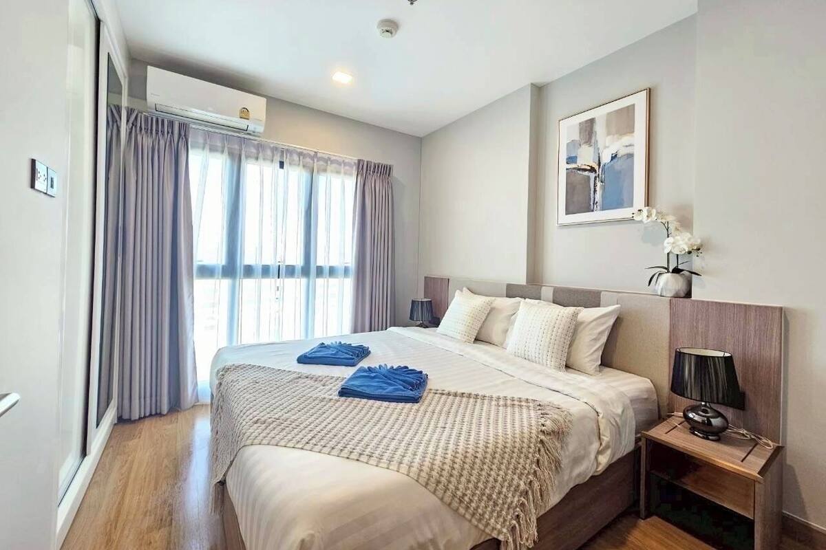 A hotel room with a large bed, a bedside table with a lamp, a wall-mounted air conditioner, and a window with curtains.