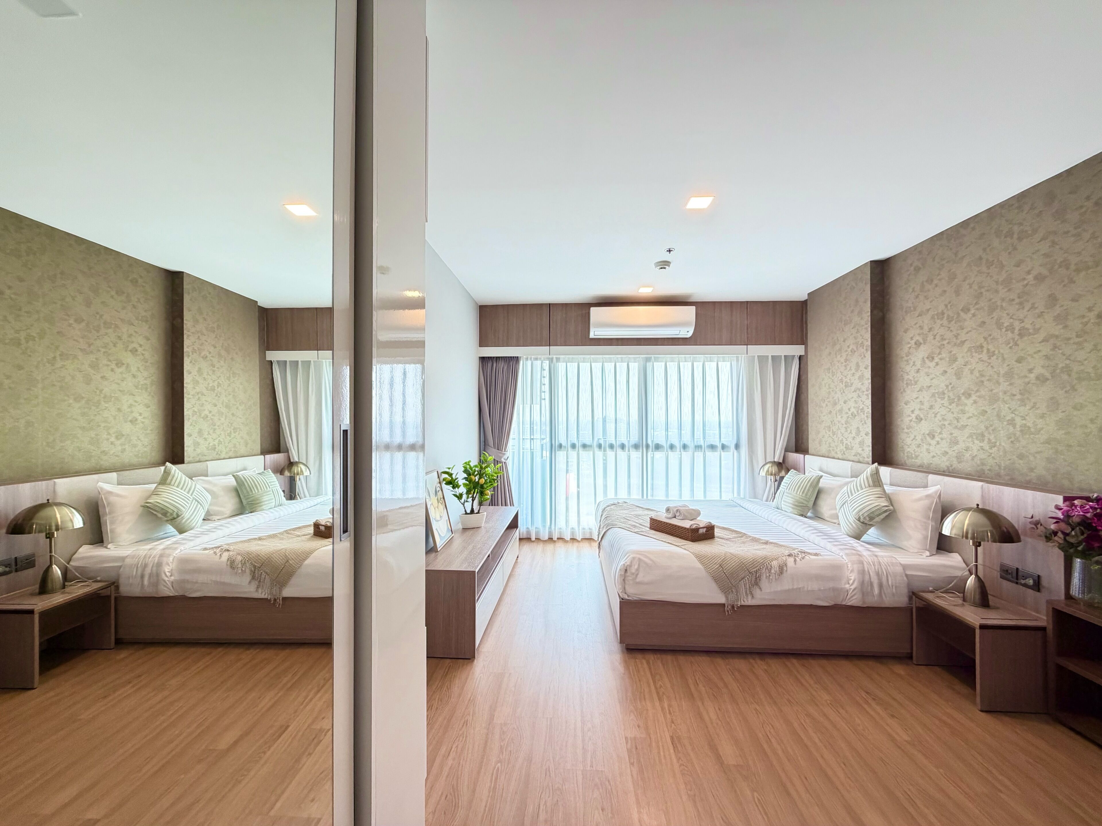 A modern hotel room with two beds, a large window with curtains, a small table with a plant, and a wall-mounted air conditioning unit.