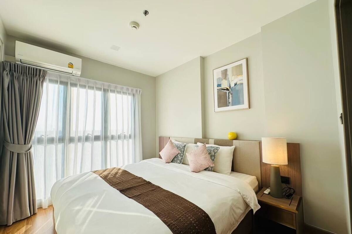 A hotel room with a large bed, a bedside table with a lamp, a window with curtains, and a ceiling light.