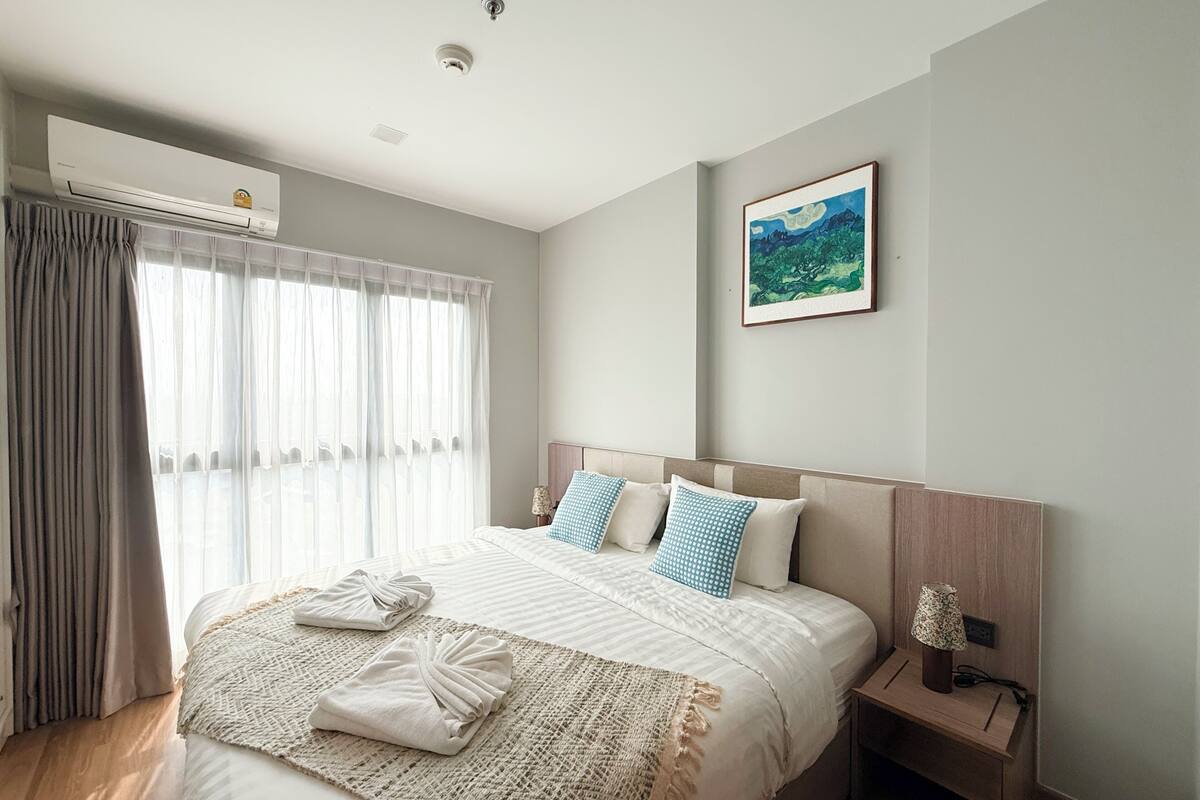 A hotel room with a bed, a nightstand, a lamp, a painting, and an air conditioning unit.
