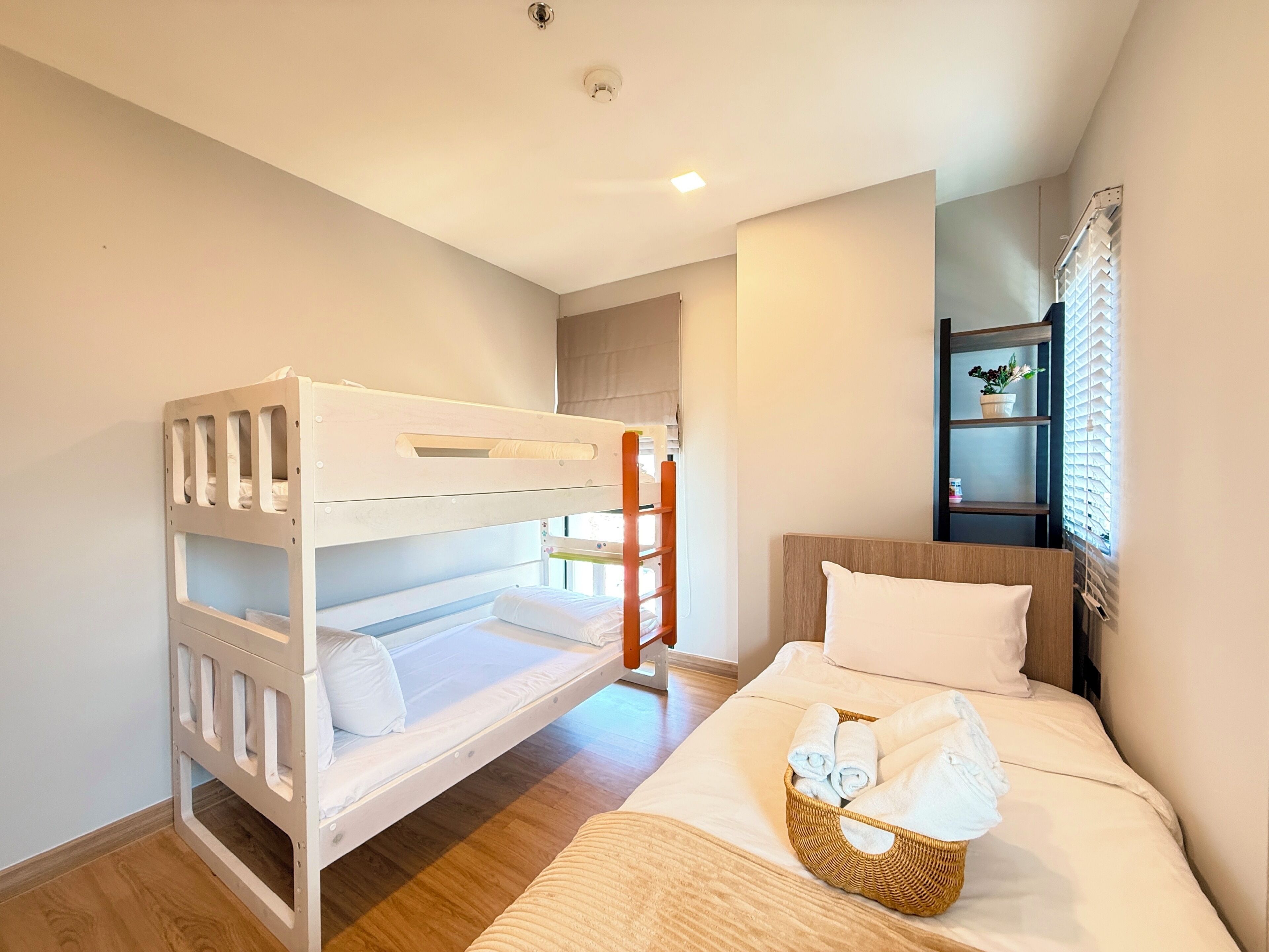 Una habitación con una cama litera, una cama individual, una cesta de mimbre con toallas, suelo de madera, una ventana con persianas y un estante con una planta.