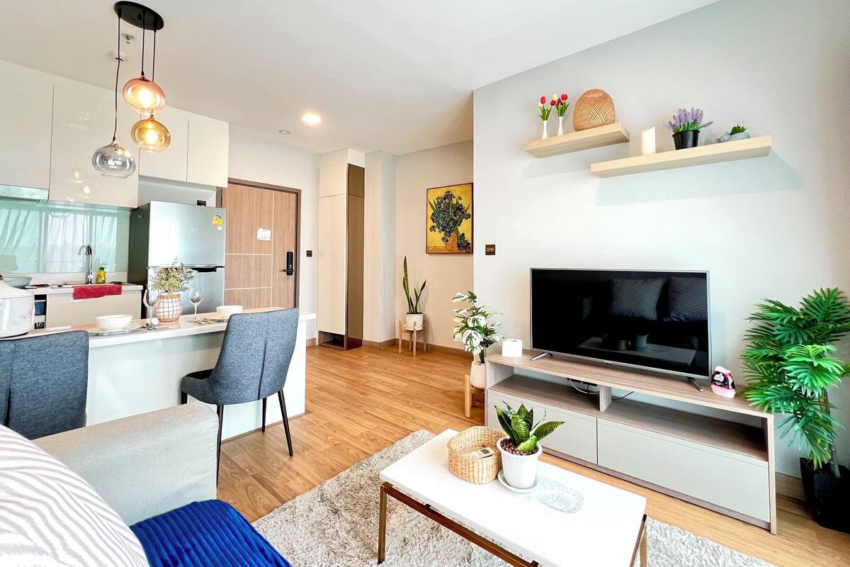 A modern living room with a TV, sofa, coffee table, and a kitchen area with a fridge and sink.
