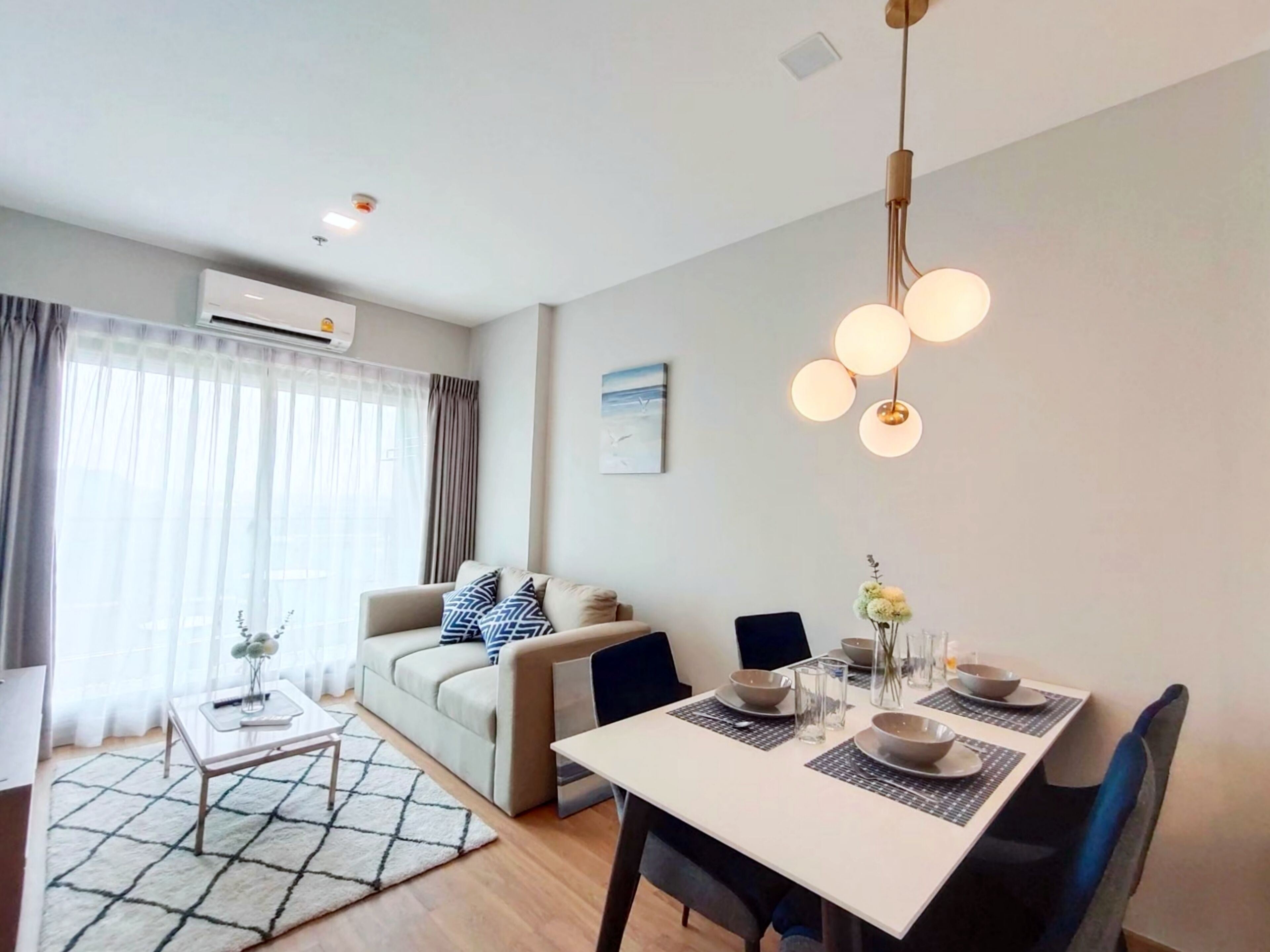 En moderne stue med sofa, spisebord og stole. Der er et vindue med gardiner, et billede på væggen og en hængende lampe.
