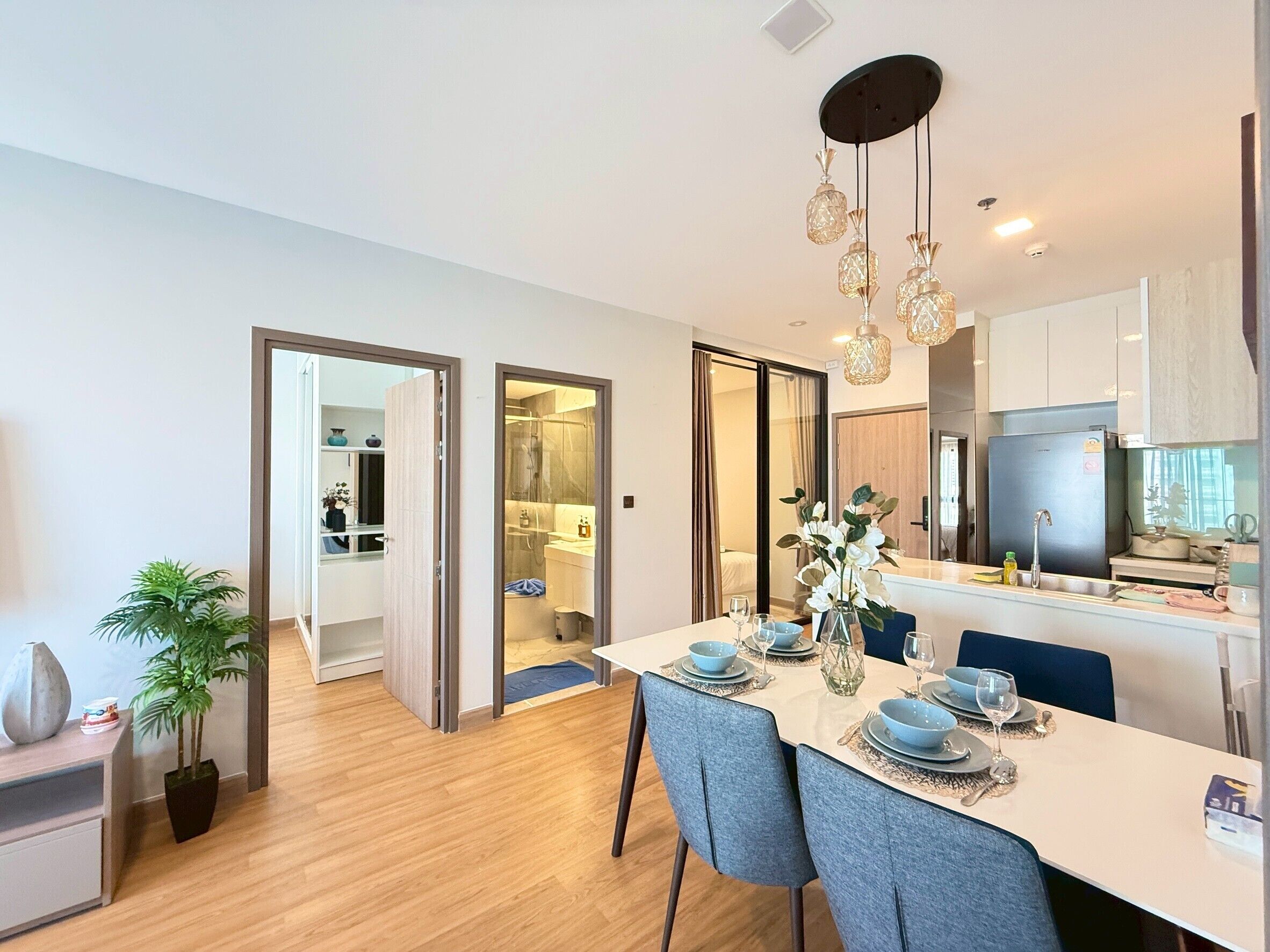 A modern kitchen with a dining area, featuring a white table set for four, a hanging light fixture, and a view into the adjacent bathroom.