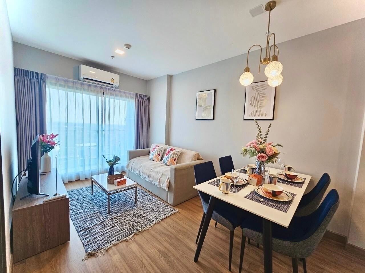 A modern living room with a sofa, dining table, and chairs. There is a TV on a stand and a window with curtains.