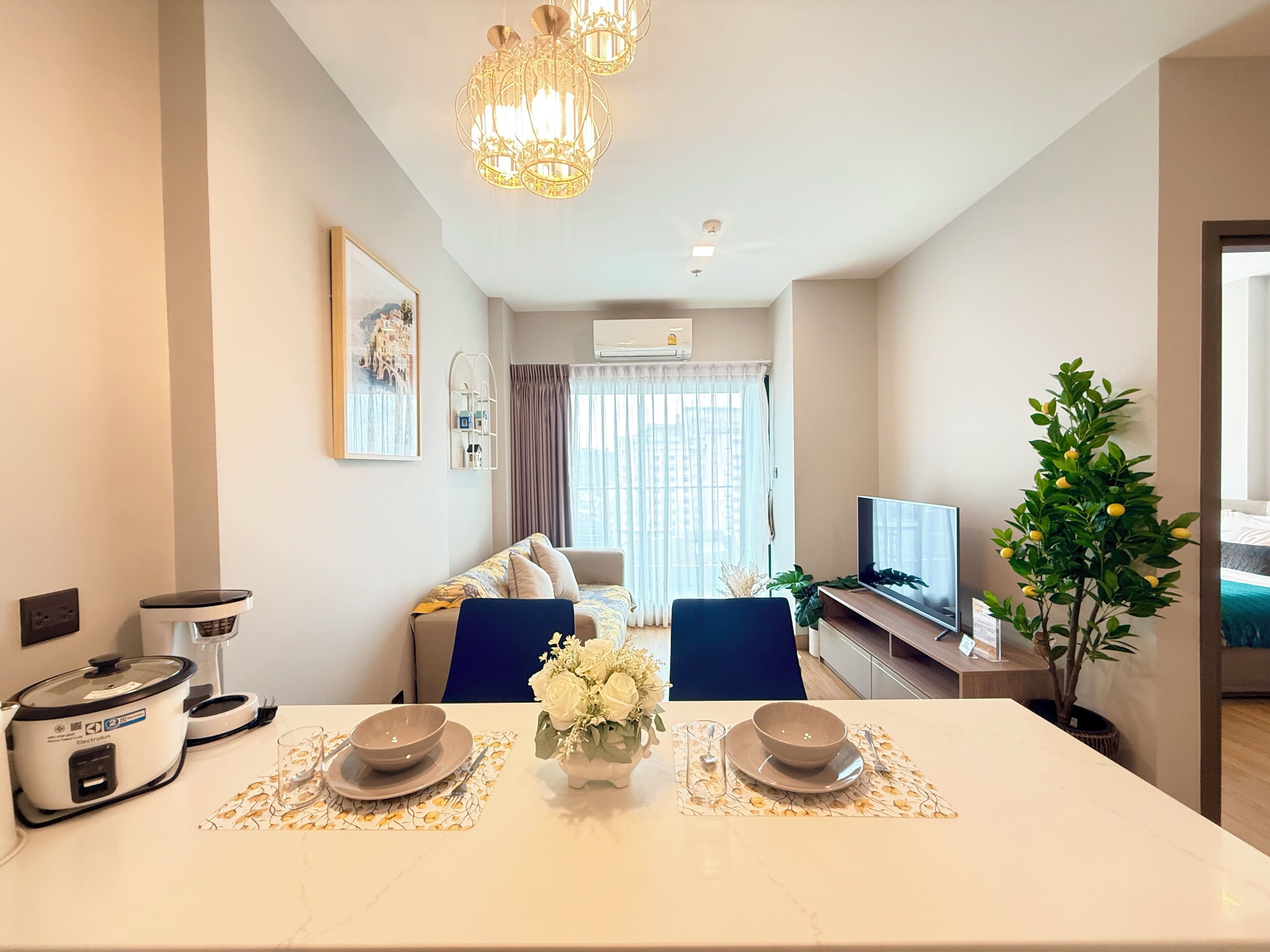 A modern living room with a dining area, a flat-screen TV, a potted plant, and a large window with curtains.