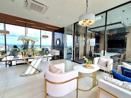 A modern hotel room with a large window offering a city view, a round table, and a seating area with white and gold furnishings.